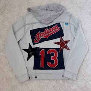 May include: Light blue denim jacket with a gray hood. Features patches with the word "Indians" in red script, the number "13" in red, and two star-shaped patches in black and red plaid.