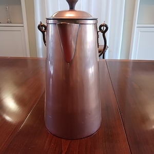 May include: A tall, copper-colored coffee pot with a dark wooden handle on the lid and black handles on the sides. The pot has a spout and a rounded base. The pot is sitting on a dark wooden table.