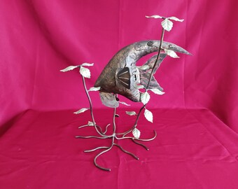 Pode incluir: Escultura em metal de um peixe com corpo texturizado cor de bronze e recortes decorativos. O peixe é sustentado por uma base de metal com caules e folhas, sobre um fundo vermelho.