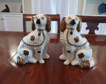 May include: Two white porcelain dog figurines with gold accents. The dogs are sitting with their heads turned to the side. They have black eyes and noses, and their ears are floppy.