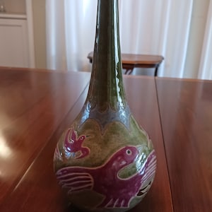 Peut inclure: Un vase en céramique vert avec un long col et un corps arrondi. Le vase est décoré de deux oiseaux dans des tons de violet et de rose.