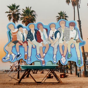 May include: A sticker of seven cartoon figures sitting on a bench. The figures are wearing colorful clothing and have different hairstyles. The sticker is placed on a lifeguard stand on a beach with palm trees in the background.