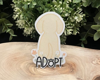 以下が含まれることがあります： 黄色い犬のステッカーで、「ADÖPT」という文字が黒で書かれています。犬は小さな花に囲まれています。ステッカーは木製の表面に貼られています。