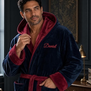 May include: A plush navy blue bathrobe featuring a deep burgundy interior lining, hood, and cuffs. The robe includes a matching burgundy tie belt and the name Daniel embroidered in red script on the left chest area of the soft fabric.