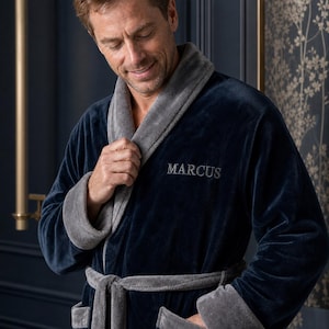 May include: A navy blue plush robe featuring a contrasting grey shawl collar, grey sleeve cuffs, and a matching grey tie belt. The name MARCUS is embroidered in grey thread on the left chest area of the soft, textured fabric garment.