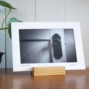 May include: Black and white framed photograph of a door with a vintage handle. The photograph is displayed on a wooden stand. A potted plant with large green leaves is in the background.