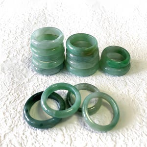 May include: A collection of jade rings in shades of green are displayed on a white, textured background. Some rings are stacked, others are scattered, highlighting the natural colour variations and polished finish. The rings vary in tone from light to dark green.