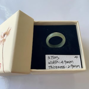 May include: A pale green jade ring displayed in an open beige box with a floral design. The ring is set against a black background. The handwritten label indicates a width of 4.9mm and a thickness of 2.9mm.