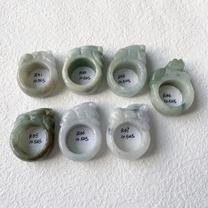 May include: Seven jade rings with carved dragon designs. The rings are in shades of green and white, with varying translucency. Each ring has a label with the letter "R" followed by a number and the measurement "10.5US".