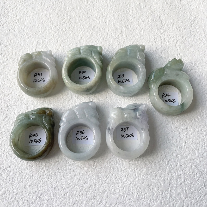 May include: Seven jade rings with carved dragon designs. The rings are in shades of green and white, with varying translucency. Each ring has a label with the letter "R" followed by a number and the measurement "10.5US".