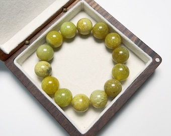 May include: A bracelet of round, light green beads with yellow and brown variations, displayed in a wooden jewelry box with a white interior. The beads are approximately 0.75 inches in diameter.