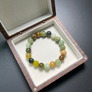 Puede incluir: Una pulsera de cuentas con tonos tierra, incluyendo verdes, marrones y amarillos. La pulsera se muestra dentro de una caja de joyería de madera con interior blanco.