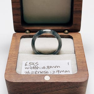 Puede incluir: Un anillo gris oscuro con apariencia veteada, exhibido en una caja de madera abierta con interior claro. El ancho del anillo es de 4,8 mm y el grosor es de 2,9 cm. El anillo está sobre un fondo blanco.