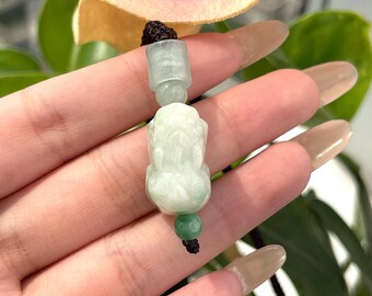 Puede incluir: Un colgante de jade verde con una figura tallada en jade de una criatura mítica. El colgante está encordado en un cordón negro con cuentas de jade verde.