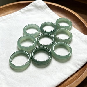 May include: A collection of eight smooth, polished, light green jade rings arranged in a cluster. Each ring features a simple, rounded band design with a translucent appearance and a consistent pale green colour throughout the set of jewellery.