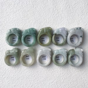 May include: A collection of ten jade rings, each carved with a detailed lion design. The rings vary in color, including shades of green, white, and lavender. Each ring has a small white tag with text. The rings are displayed on a white surface.
