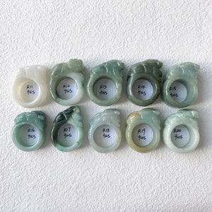 May include: A collection of ten jade rings in varying shades of green and white. Each ring features a carved design and a label with a number and the letters "qus". The rings are displayed on a white surface.