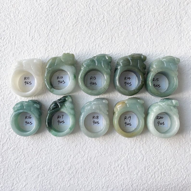 May include: A collection of ten jade rings in varying shades of green and white. Each ring features a carved design and a label with a number and the letters "qus". The rings are displayed on a white surface.