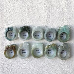 May include: Ten jade rings with carved animal designs are displayed. The rings vary in color, including green, white, and brown. Each ring has a tag with the text "R24 9.5us". The rings are arranged on a white surface.
