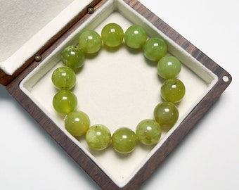 May include: A beaded bracelet composed of round, light green beads. The bracelet is displayed inside a wooden box with a white, cushioned interior. The beads have a translucent quality and varying shades of green.