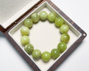 May include: A bracelet of large, round, light green beads rests inside a wooden jewelry box with a white interior. The beads have a smooth, polished appearance and vary slightly in color and pattern. The box is open, revealing the bracelet's full circular form.