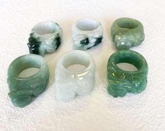 May include: Six jade rings in shades of green and white are shown. Each ring has a carved design and a smooth, polished finish. The rings are arranged on a white surface, highlighting their intricate details and natural colours. The rings are likely made of jade.
