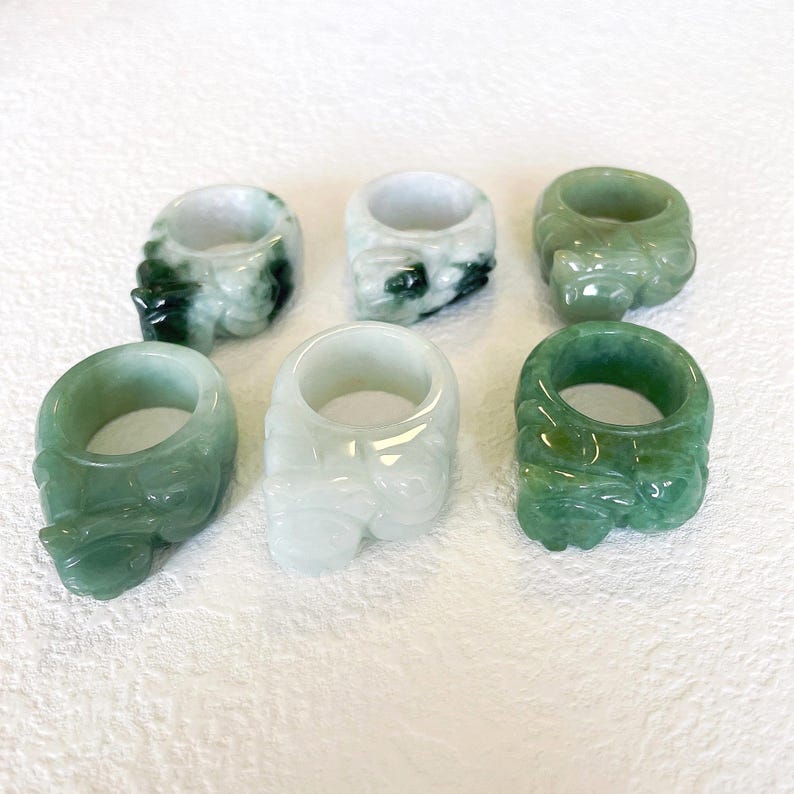 May include: Six jade rings in varying shades of green and white are displayed. Each ring features a carved design and a smooth, polished finish. The rings are arranged on a white surface, showcasing their intricate details and natural colors.