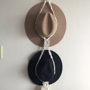 May include: White macrame hat rack with two hats hanging on it. The top hat is brown and the bottom hat is black.