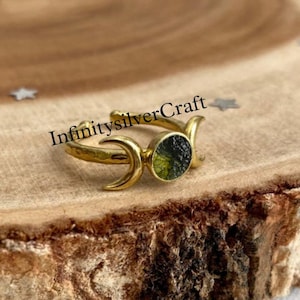 May include: A gold-colored ring with a green stone center, flanked by crescent moon designs. The ring is set on a wooden surface with star-shaped confetti. The text "InfinitySilverCraft" is visible.