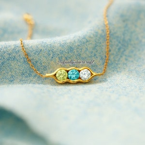 Op de afbeelding: Een goudkleurige ketting met een erwtpeul-hanger met drie ronde edelstenen in lichtgroen, turquoise en wit. De ketting ligt op een blauwe stoffen achtergrond.