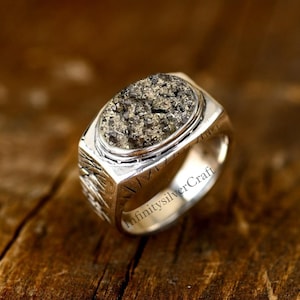 Puede incluir: Un anillo de plata con una piedra de pirita de forma ovalada engastada en un engaste de bisel. La banda del anillo es texturizada y tiene las palabras "InfinitysilverCraft" grabadas en el lateral.