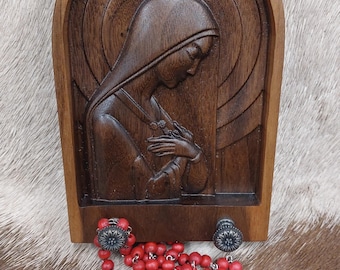 May include: A wooden wall plaque with a carved image of the Virgin Mary. The plaque has a rounded arch top and is decorated with a circular pattern. A red rosary with a silver cross is lying in front of the plaque.