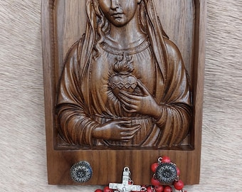 May include: A wooden wall hanging featuring a carved image of a religious figure holding a flaming heart. Below, a red beaded rosary with a silver crucifix hangs. The wall hanging has two decorative knobs.