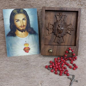 May include: A religious set featuring a portrait of Jesus Christ, a wooden box with a carved cross design, and a red beaded rosary with a silver crucifix. The items are arranged on a textured, light brown surface.