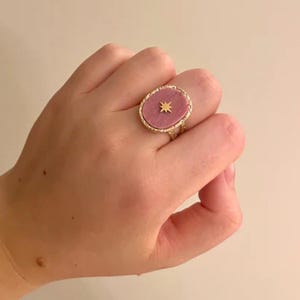 Op de afbeelding: Een goudkleurige ring met een ovale, roze steen en een gouden sterrenpatroon. De ring heeft een getextureerde, sierlijke band. De ring wordt aan een vinger gedragen.