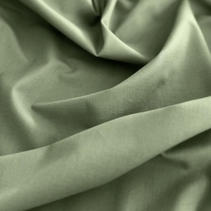 Könnte beinhalten: Nahaufnahme eines olivgrünen Stoffes mit sanften Falten und feiner Textur. Das Material scheint glatt, möglicherweise gewebt, mit verschiedenen Grüntönen, die durch die Falten und das Licht entstehen. Der Gesamteindruck ist von Weichheit und Tiefe geprägt.