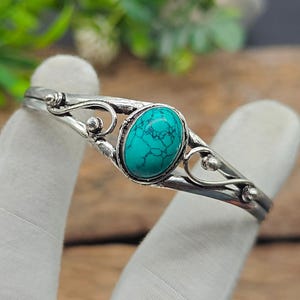 May include: A silver-toned bracelet featuring an oval turquoise stone. The bracelet has an open design with decorative swirls and small bead accents. The turquoise stone is a vibrant blue-green color, adding a pop of color to the piece.