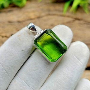 Puede incluir: Colgante rectangular de piedra preciosa verde brillante con engaste plateado. El colgante se sostiene sobre una superficie blanca, con fondo de madera y follaje verde visible.