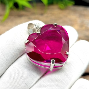 Könnte beinhalten: Ein herzförmiger Anhänger mit einem facettierten, tiefrosa Edelstein. Der Edelstein ist in einem silberfarbenen Metallrahmen mit einer Öse oben für eine Kette gefasst. Der Anhänger wird von einer behandschuhten Hand gehalten.