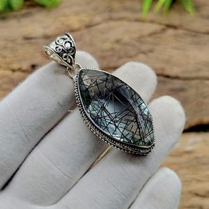 May include: A silver pendant with an oval, clear gemstone featuring black inclusions. The gemstone is set in a detailed silver frame with a decorative bail. The pendant is held in a gloved hand.