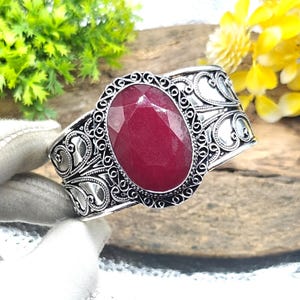 May include: A silver cuff bracelet featuring an oval-shaped, faceted deep red gemstone at the center. The metal band displays intricate filigree scrollwork and a beaded border surrounding the central stone, creating a detailed metallic texture.