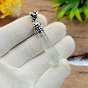 May include: A clear, faceted crystal pendant with a silver-coloured metal setting. The pendant is elongated and has a pointed tip. The metal setting features decorative swirls and a small loop for a chain. The crystal is approximately 5 cm long.