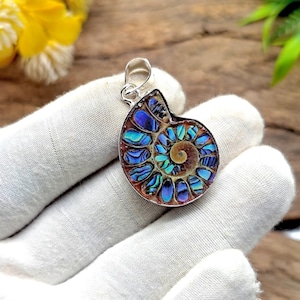 Pode incluir: Pingente de prata com um fóssil de amonite iridescente. O fóssil apresenta um padrão espiral com tons de azul, verde-azulado e castanho. O pingente é segurado por uma mão enluvada, destacando os detalhes intrincados.