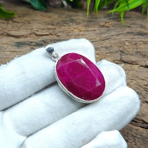 May include: A vibrant ruby-red oval pendant with a faceted surface, set in a silver-toned metal frame. The pendant is displayed against a white fabric background, highlighting the gemstone's colour and cut. The pendant has a small loop at the top for a chain.