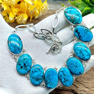 May include: A silver bracelet featuring oval-shaped turquoise stones. The bright blue stones are set in a silver chain, creating a striking contrast. The bracelet is displayed on a white surface, highlighting the jewelry's design.