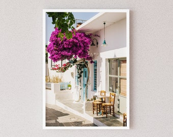 May include: A white building with a blue door and a pink bougainvillea bush growing on the side. There are two wooden chairs and a small table in front of the building.