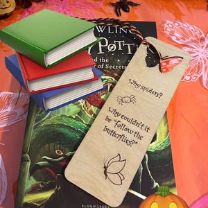 May include: A stack of three miniature books in green, red, and blue, resting on a Harry Potter and the Chamber of Secrets book. A light-coloured wooden bookmark features a butterfly, a spider icon, and the text: Why spiders? Why couldn't it be follow the butterflies?