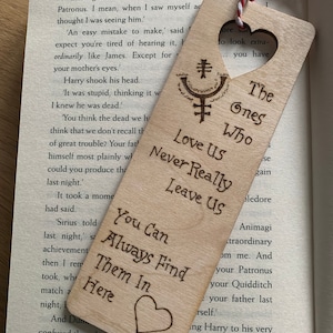 May include: Wooden bookmark with a heart shape and the text "The Ones Who Love Us Never Really Leave Us You Can Always Find Them In Here". The bookmark also features a symbol with a circle and a cross.