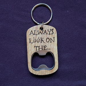 May include: A wooden bottle opener with a silver metal opener and a keyring. The opener is engraved with the words "ALWAYS LOOK ON THE..." in a serif font. The wood has a light brown colour.