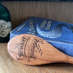 May include: A wooden spoon with the words "Mischief Managed" engraved on it. The spoon is resting on a blue book titled "Advanced Potion Making".